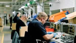 Worker in a factory sits at a bench assembling small parts with blue gloves, orange bins and other workers visible in the background.